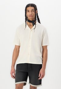 Short-sleeve, cream knit shirt with a classic collar, button front, and textured pattern. Worn with dark denim shorts and a black scarf.