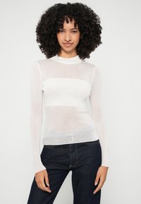 Bela rebrasta majica z dolgimi rokavi in visokim ovratnikom, ki ima polprosojno teksturo in vodoravne črte. Kombinirana z temnimi džinsom.