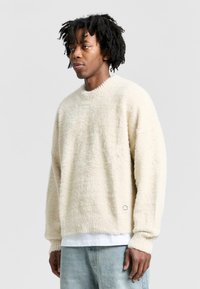 Cozy cream sweater made of fluffy material, featuring a loose fit, ribbed cuffs, and a subtle logo at the hem. Paired with light jeans.