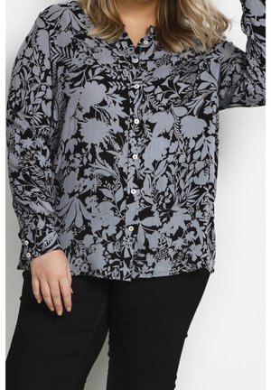 Femme portant une chemise à manches longues boutonnée avec un motif floral gris sur tissu noir et un pantalon noir, debout, main près des cheveux.