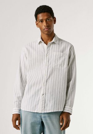 Junger Mann trägt ein weißes, langärmliges Hemd mit dünnen vertikalen Streifen und hellblaue Jeans, steht vor einem einfarbigen Hintergrund.