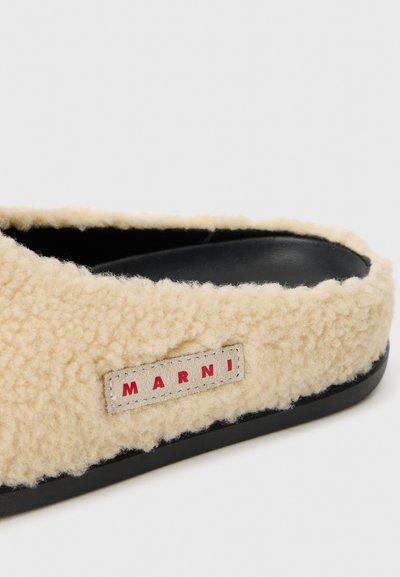 Beige fluffig slip-on sandal med övre av shearling, svart innersula och en grå etikett med röd "MARNI" text. Platt svart sula.
