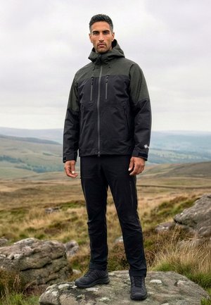 Mand står på en stor sten iført sort hættejakke til udendørs brug, sorte bukser og vandresko med græsklædte bakker og en skyet himmel i baggrunden.