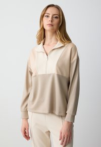 Beige zip-up sweatshirt met een opstaande kraag, met een kleurblokkenschema en een zachte, gladde textuur. Gecombineerd met lichtbeige broeken.