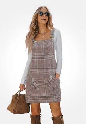 Vestido peto a cuadros grises con tirantes ajustables, dos bolsillos delanteros y escote cuadrado. Estilizado con una blusa de manga larga y un bolso marrón.