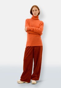 Orange Rollkragenpullover aus weichem Strick, kombiniert mit weitgeschnittenen, rostfarbenen Cordhosen, die eine strukturierte Oberfläche und gerippte Bündchen aufweisen.