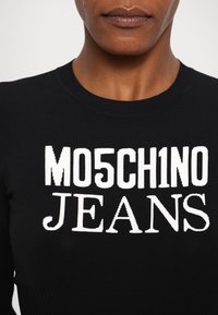 Sweater preto com nervuras, apresentando letras brancas em destaque "MO5CH1NO JEANS." O design destaca um decote redondo e uma forma ajustada.