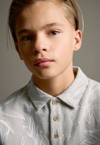 Hemd mit Kragen aus hellgrauem Stoff mit Blattmuster, Knopfleiste vorne und strukturierter, gewebter Oberfläche.