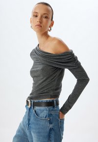 Graues, schulterfreies Oberteil mit langen Ärmeln und gerafftem Stoff, kombiniert mit hoch taillierten, ausgewaschenen blauen Jeans und einem schwarzen Gürtel mit einer silbernen Schnalle.