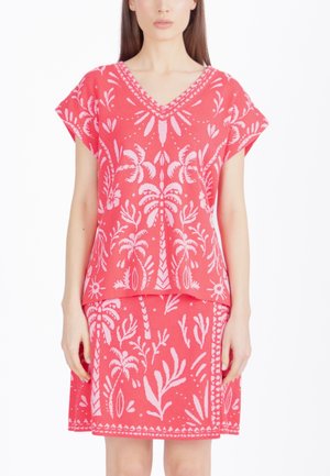 Femme portant un ensemble haut et jupe corail avec des motifs légers de feuilles et palmiers tropicaux rose pâle, debout devant un fond blanc.