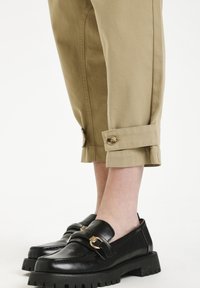 Kaki cropped byxor med uppvikta muddar och knappdetalj, i kombination med svarta läderloafer med guldhardware och kraftiga sulor.