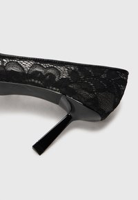 Pantofi cu toc din dantelă neagră, cu vârf ascuțit, toc stiletto lucios negru și detalii cu overlay din dantelă texturată. Siluetă elegant curbată.