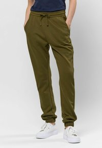 Olijfgroene joggers van zachte stof met een elastische tailleband en een trekkoord, voorzien van zijzakken en elastische boorden. Gepaarde met witte sneakers.