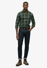 Groen en blauw geruite shirt, denim jeans, bruine veterschoenen, riem, model staat met handen in de zakken, casual pasvorm, opgerolde jeans cuffs.
