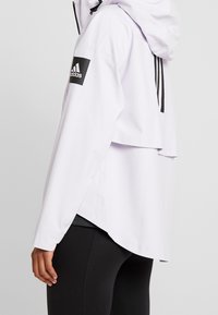 Vista lateral de una persona que lleva un hoodie blanco de Adidas con el logo en la manga y leggings negros, contra un fondo liso.