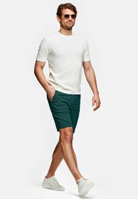 Man loopt met een zonnebril, een wit T-shirt met korte mouwen, donkergroene korte broek en witte sneakers tegen een effen witte achtergrond.