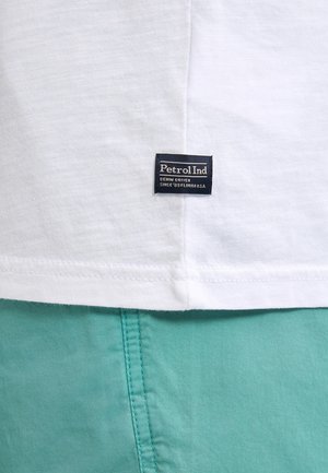 Weißes Baumwoll-T-Shirt mit einem blauen rechteckigen Etikett mit der Aufschrift "Petrol Ind" nahe dem Saum. Teal-farbene Baumwollshorts mit sichtbaren Nahtdetails.