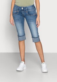 Světle modré denimové capri džíny s ohrnutými nohavicemi, pěti kapsami a detaily v oranžovém šití, spárované s bílými teniskami na neutrálním pozadí.