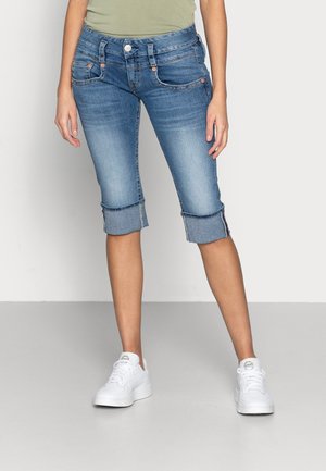 Pantalones capri de mezclilla azul claro con puños enrollados, cinco bolsillos y detalles de costura en naranja, combinados con zapatillas blancas sobre un fondo neutro.