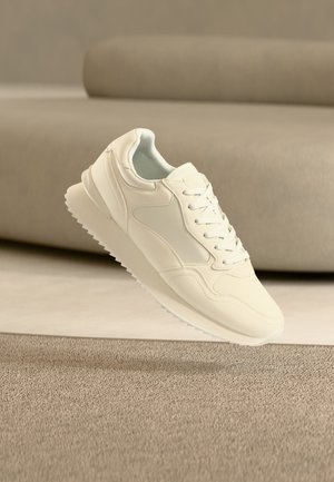 Baskets de couleur crème avec un design à lacets, une semelle épaisse et une semelle extérieure texturée, flottant au-dessus d'un tapis beige et d'un canapé en arrière-plan.