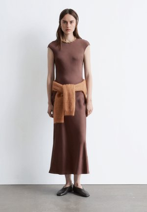 Braunes, satineng anliegendes Kleid mit kurzen Ärmeln und knielangem Saum, dazu ein flauschiger, oranger Pullover, der in der Taille gebunden ist. Modell trägt dunkle Slipper.