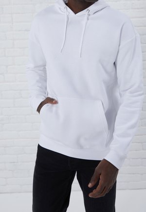 Witte hoodie van zachte stof, met een voorzak, een capuchon met trekkoord en een geribbelde zoom. Gekocht met zwarte broek, voor een bakstenen muur.