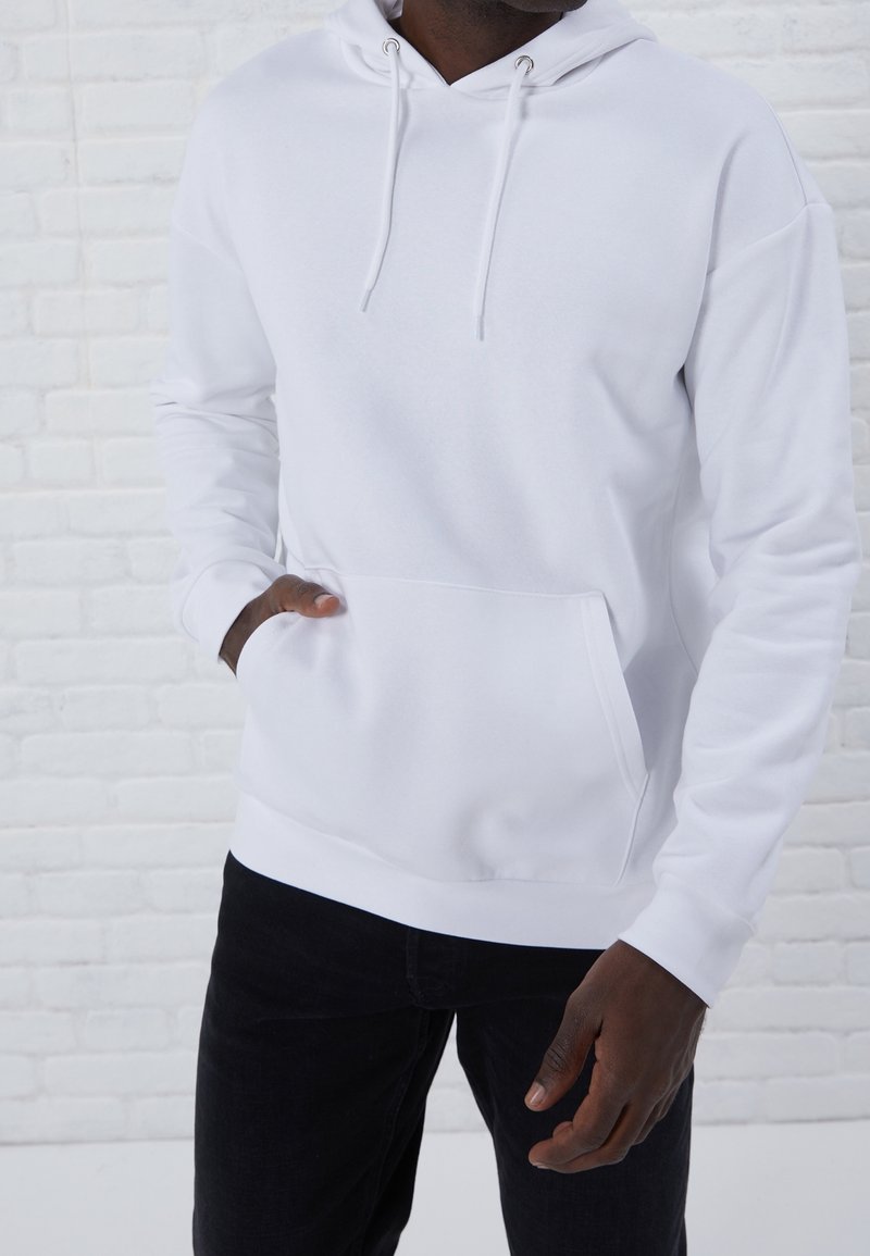 Sudadera blanca de tejido suave, con un bolsillo delantero, capucha con cordón y dobladillo acanalado. Usada con pantalones negros, frente a una pared de ladrillos.