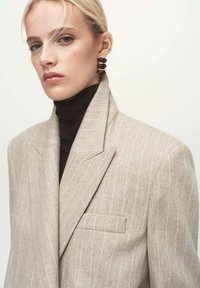 Blazer de rayas beige con solapas afiladas, forma estructurada y un bolsillo frontal; llevado sobre un cuello de tortuga negro. Aretes en espiral dorados como detalle.