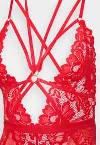 Rød blonde bodystocking med blomsterprint, justerbare stropper og en cirkulær guld-ringdetalje på ryggen. Gennemsigtig tekstur og krydsdesign.