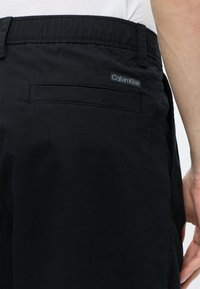 Μαύρο παντελόνι Calvin Klein, εμφανιζόμενο από πίσω στη μέση, με τσέπη welt και ορατή ετικέτα του brand σε άτομο που φοράει λευκό πουκάμισο.