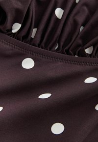 Tissu marron à pois blancs, avec un détail froncé le long du bord supérieur. Texture douce avec un léger éclat, ourlet soigneusement cousu.