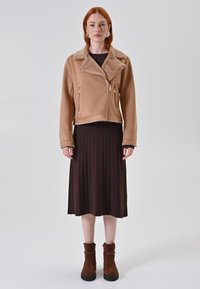 Giacca beige in finta pelliccia con colletto a rever, tasche con zip e vestibilità corta, abbinata a un vestito a coste scuro e stivaletti alla caviglia marroni.