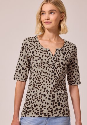 Lyshåret kvinde iført en beige t-shirt med korte ærmer med sorte uregelmæssige pletter og blå jeans, der kigger let til siden.