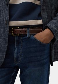 Jeans in denim blu scuro con una mano in tasca, abbinati a un maglione a righe blu navy e beige e a un blazer grigio testurizzato. Cintura in pelle marrone con fibbia in ottone.