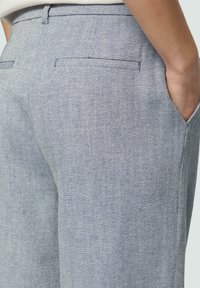 Blauwe broek met een visgraatpatroon en een gladde textuur, met achterzakken en een getailleerde pasvorm. Zichtbare stiksels langs de naden.