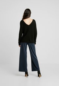 Czarny sweter o luźnym kroju z dekoltem w serek i fakturowanym splotem; w połączeniu z szerokimi, granatowymi spodniami; modelka ma na sobie czarne botki za kostkę.