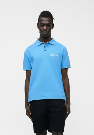 Man wearing a bright blue polo shirt with "Karl Lagerfeld Jeans" logo and black shorts, standing against a plain white background.