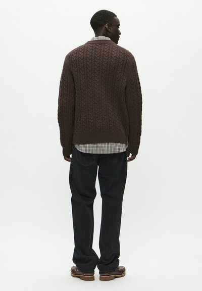 Rjava pletiva pulover, v kombinaciji s sivo karirasto srajco spodaj, temnimi jeans hlačami in rjavimi čevlji. Čista, teksturirana tkanina.