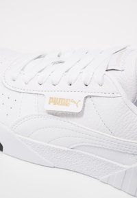 Białe skórzane sneakersy mają fakturowaną powierzchnię, białe sznurówki, perforacje oraz złotą metkę z logo. Grube białe podeszwy z czarnymi akcentami.