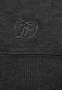 Tessuto a maglia grigio scuro con una lettera "D" stilizzata ricamata sopra una linea di cucitura testurizzata.