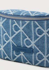 Μπλε τζιν τσάντα με γεωμετρικό λευκό σχέδιο, φερμουάρ κλεισίματος και δερμάτινη ετικέτα Calvin Klein στο πλάι. Υφασμάτινη υφή.