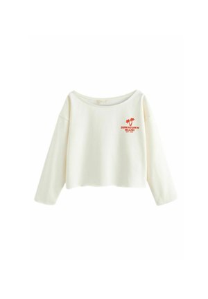 Femme portant un sweat-shirt blanc épaules dénudées avec le logo « Downtown Miami » et un short noir ajusté, debout à l'extérieur.