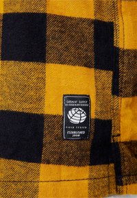 Geel en zwart geblokt stof met een geweven textuur, met een zwarte label waarop een wereldbol-logo en de tekst "FIELD TESTED ESTABLISHED 1998" staat.