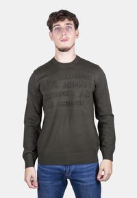 Maglione verde scuro con dettaglio del logo testurizzato sul petto, collo rotondo e polsini a costine. Indossato con jeans in denim blu.