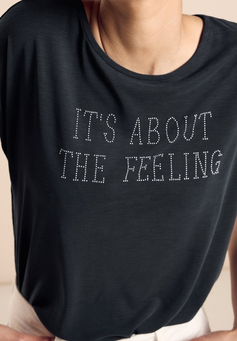 Tmavě šedé tričko s krátkým rukávem, volného střihu a kulatým výstřihem, s bílým textem ozdobeným kamínky, který zní: "JE TO O POCITU."