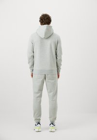 Mężczyzna odwrócony plecami, ubrany w jasnoszary hoodie i pasujące dresy, w białych sneakersach, stojący na tle czystej białej ściany.