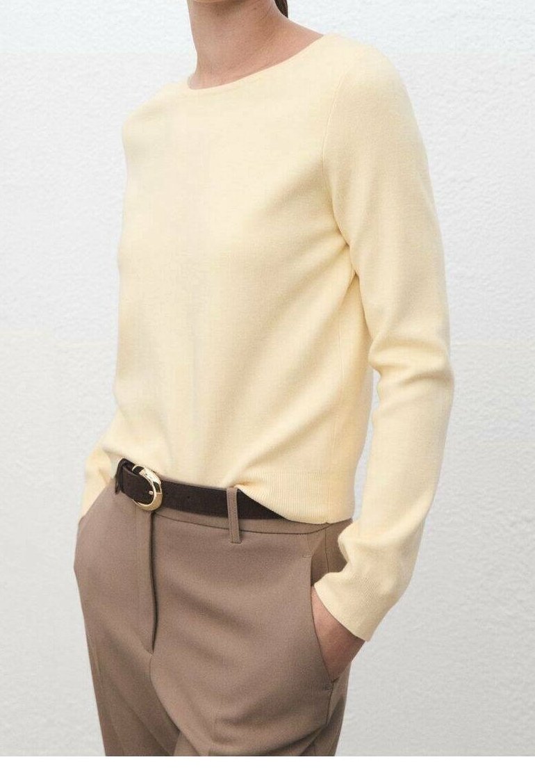 Personne portant un haut à manches longues jaune clair rentré dans un pantalon taille haute taupe avec une ceinture noire, les mains dans les poches.