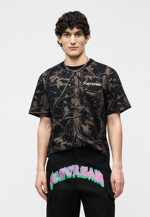 Jonge man met krullend haar, gekleed in een zwart T-shirt met camoufleringsprint en een zwarte broek met kleurrijke "ICECREAM"-tekst aan de voorkant.