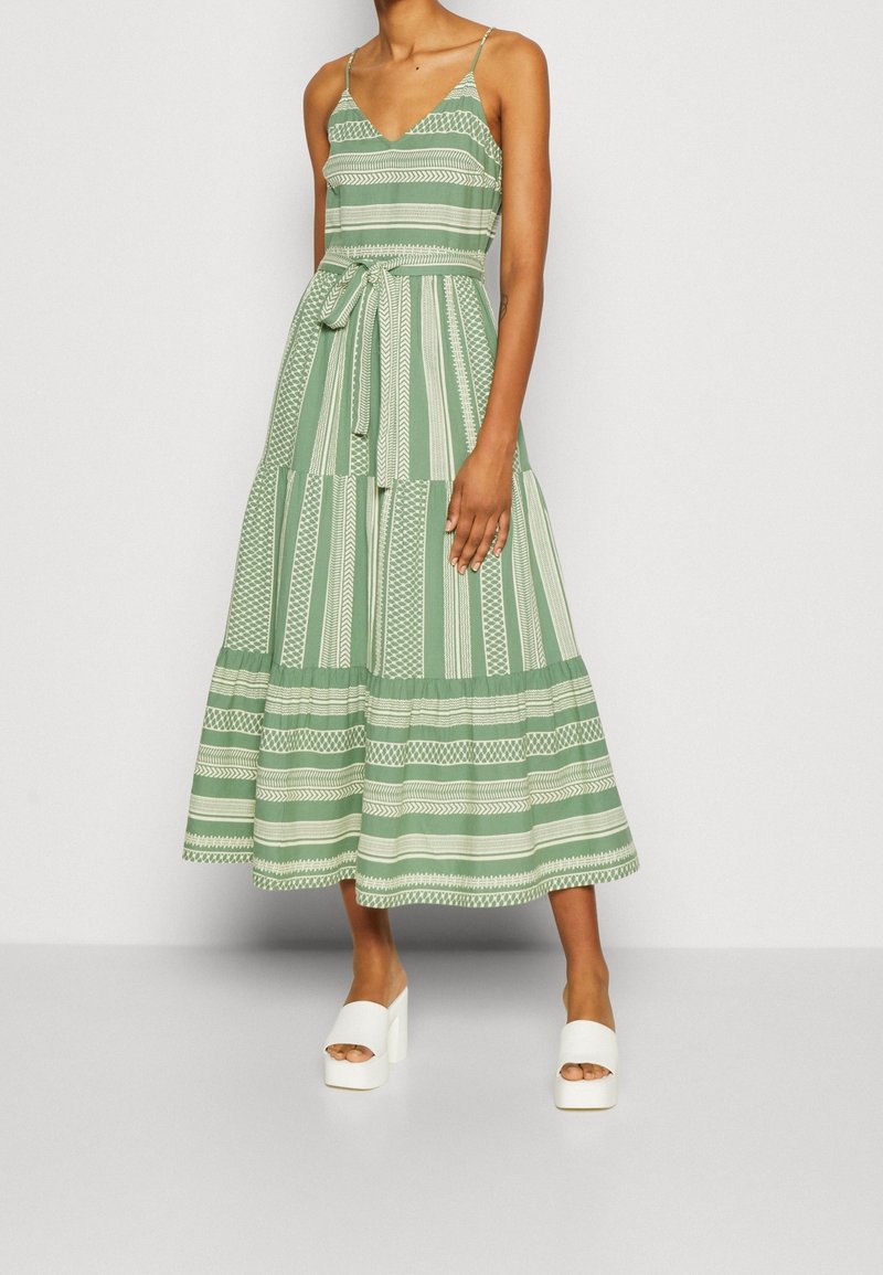 Femme portant une robe midi sans manches à motifs verts et blancs avec une ceinture nouée à la taille et des chaussures blanches à plateforme à bout ouvert sur un fond uni.