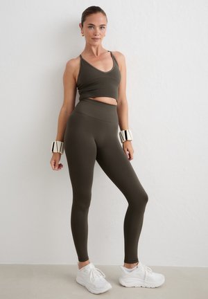 Femme portant un ensemble de sport vert foncé avec des leggings taille haute et un débardeur court, des baskets blanches et des bracelets blancs, debout contre un mur blanc.